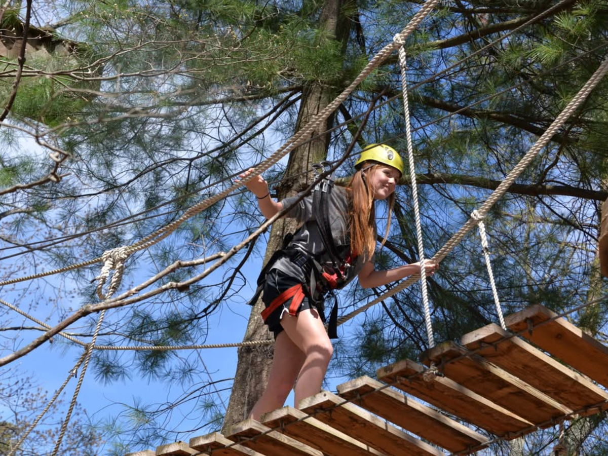 A young woman wearing a helmet and harness carefully crosses a wooden rope bridge high in the trees on an outdoor adventure course.