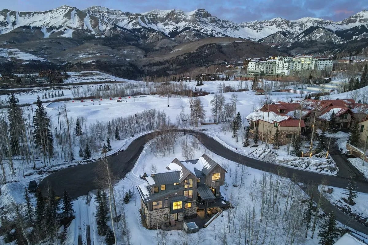 Aerial view of a modern mountain home with glowing windows, surrounded by snow-covered trees, winding roads, and ski slopes beneath dramatic alpine peaks.