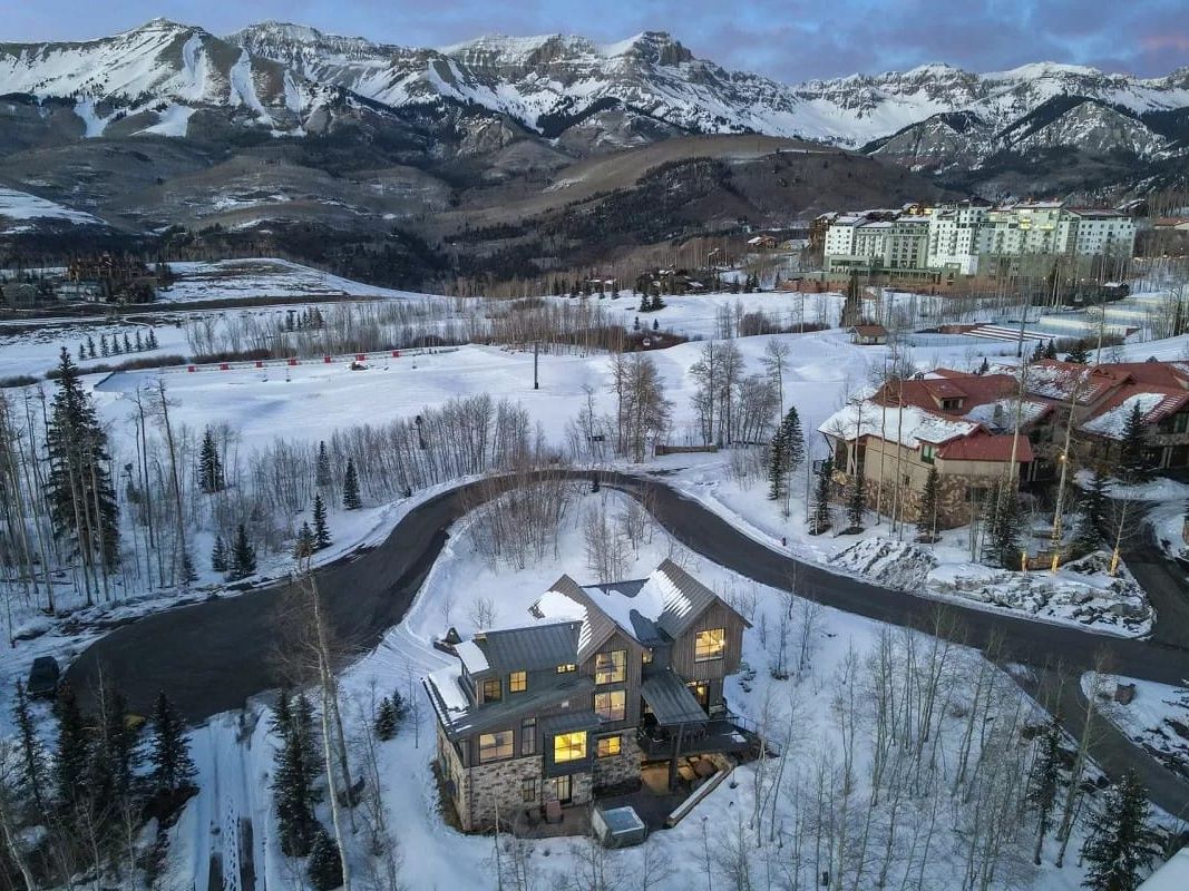 Aerial view of a modern mountain home with glowing windows, surrounded by snow-covered trees, winding roads, and ski slopes beneath dramatic alpine peaks.