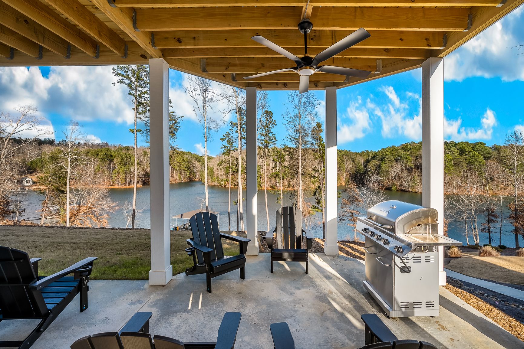 Covered patio with Adirondack chairs, a grill, and ceiling fan overlooking a peaceful lake surrounded by trees.