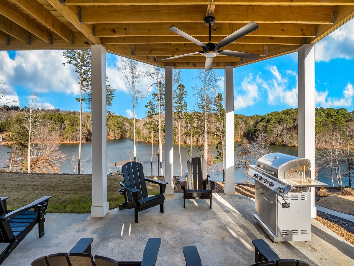 Covered patio with Adirondack chairs, a grill, and ceiling fan overlooking a peaceful lake surrounded by trees.