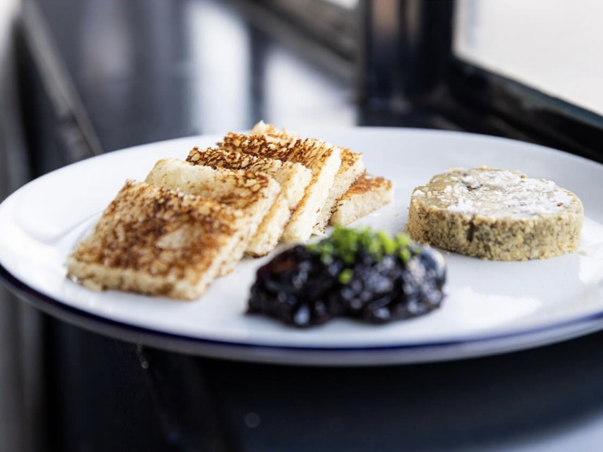 White plate with neatly stacked grilled toast triangles, a round slice of pâté, and a small mound of dark fruit chutney topped with chopped herbs.