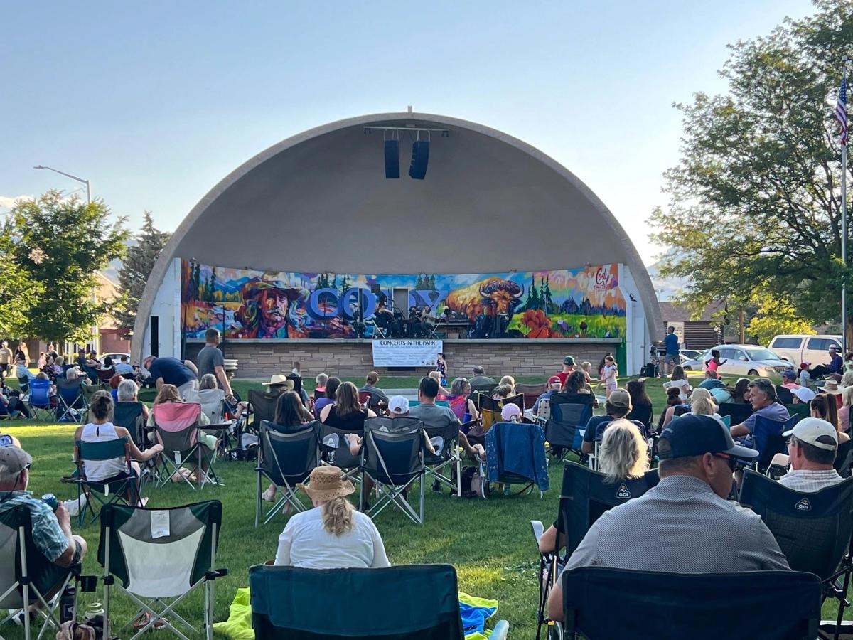 Families and friends relax on lawn chairs and blankets in front of Cody’s outdoor stage, enjoying live music under a sunny blue sky. The colorful mural behind the band adds to the cheerful small-town community vibe.