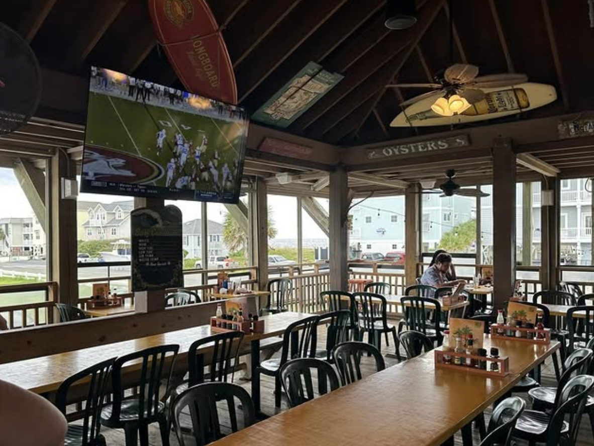 A casual beachside seafood restaurant with wooden tables, plastic chairs, surfboards on the ceiling, and a large TV showing a football game.