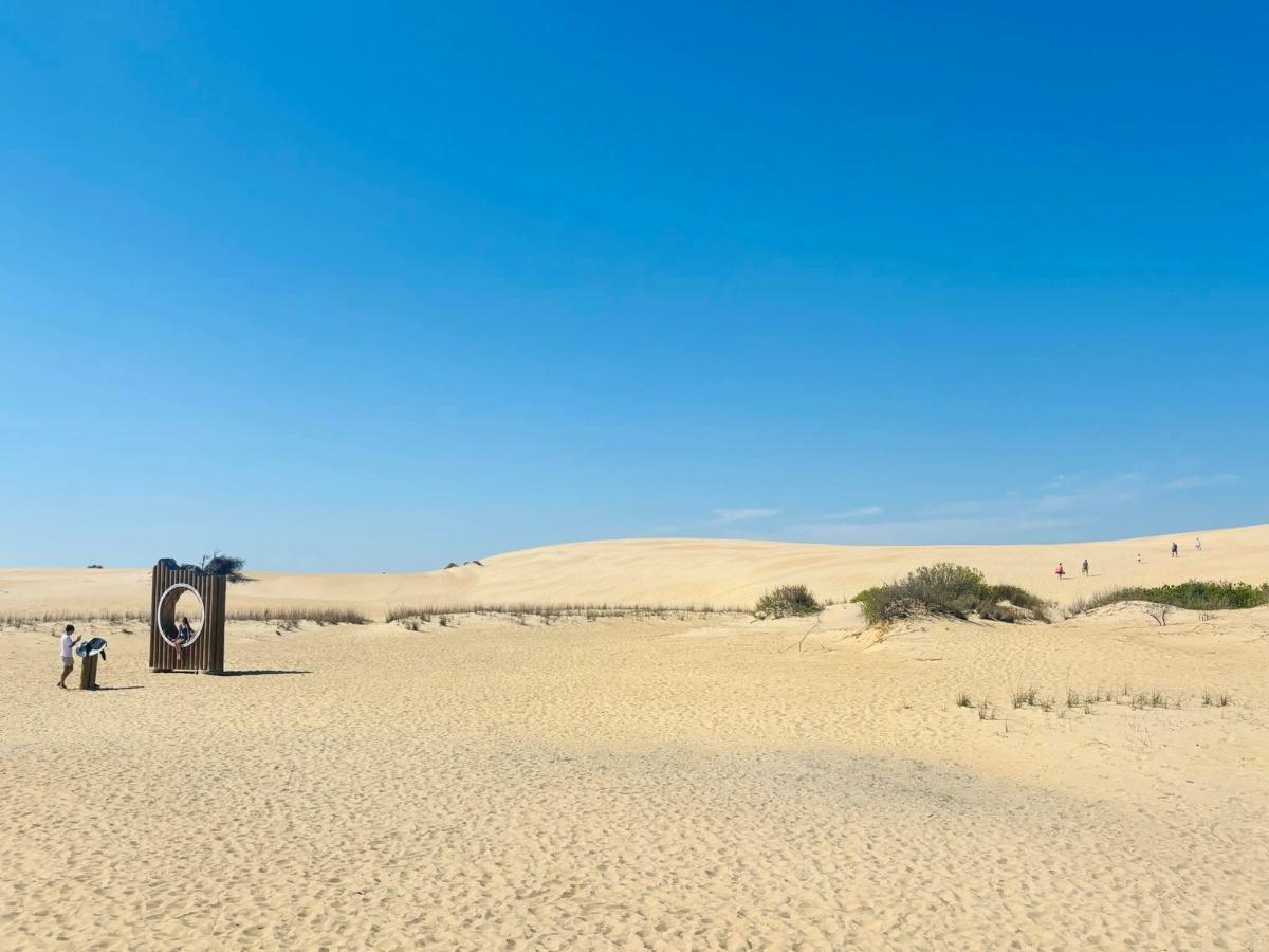 Two visitors pause at a wooden framed viewing structure at the base of the vast sand dunes of Jockey's Ridge, with tiny figures scattered across the golden slopes in the distance. A brilliant clear blue sky fills the upper half of the expansive desert-like landscape.