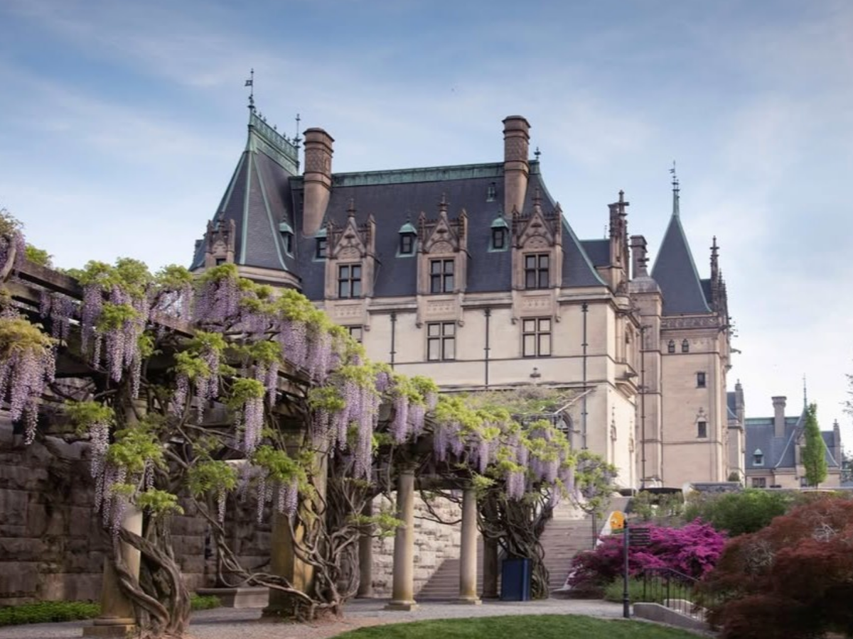 Elegant historic mansion with steep slate rooftops and ornate architecture, framed by wisteria-draped trellises and colorful spring gardens