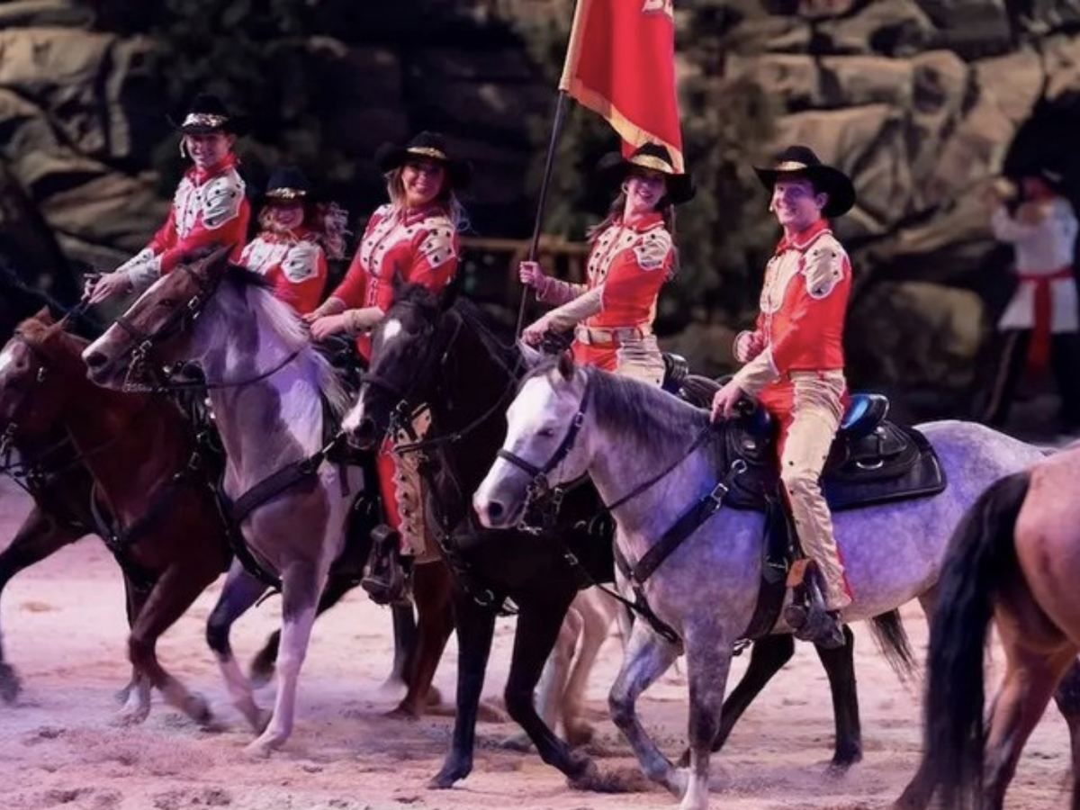 Performers in red and white costumes ride horses in an arena, one carrying a red flag, as part of a lively equestrian show with a western theme.