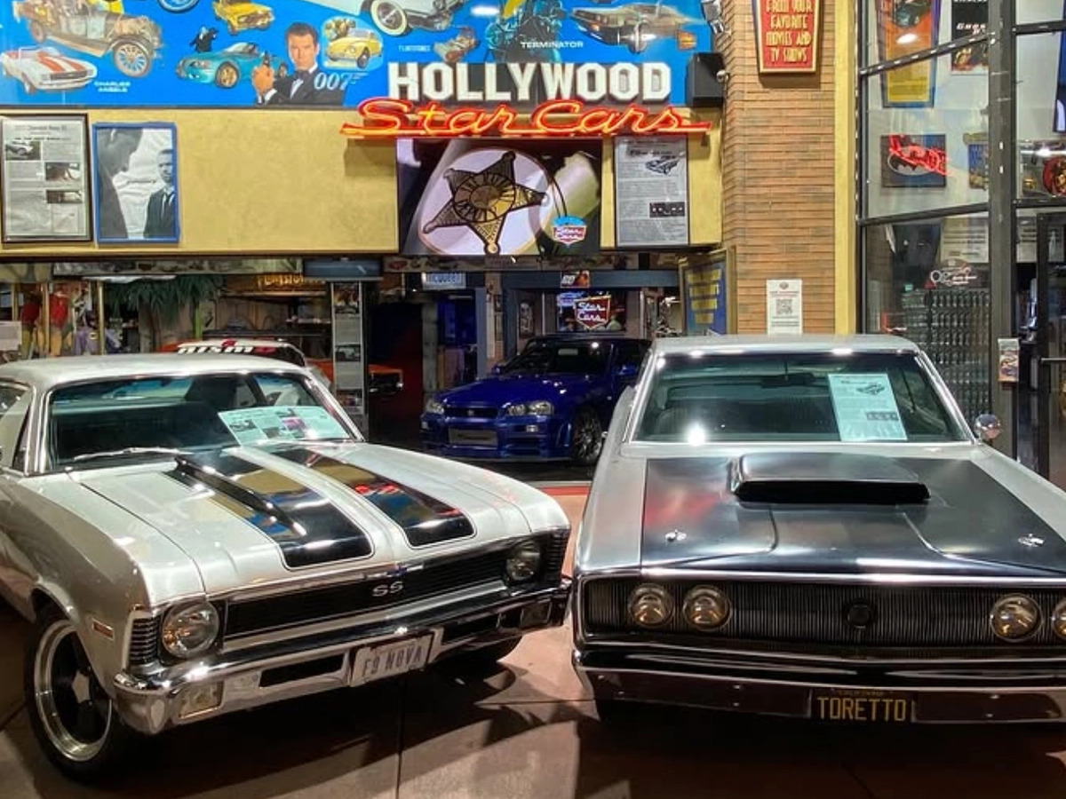 Two famous muscle cars are displayed side by side at the Hollywood Star Cars Museum, with movie posters and memorabilia in the background.