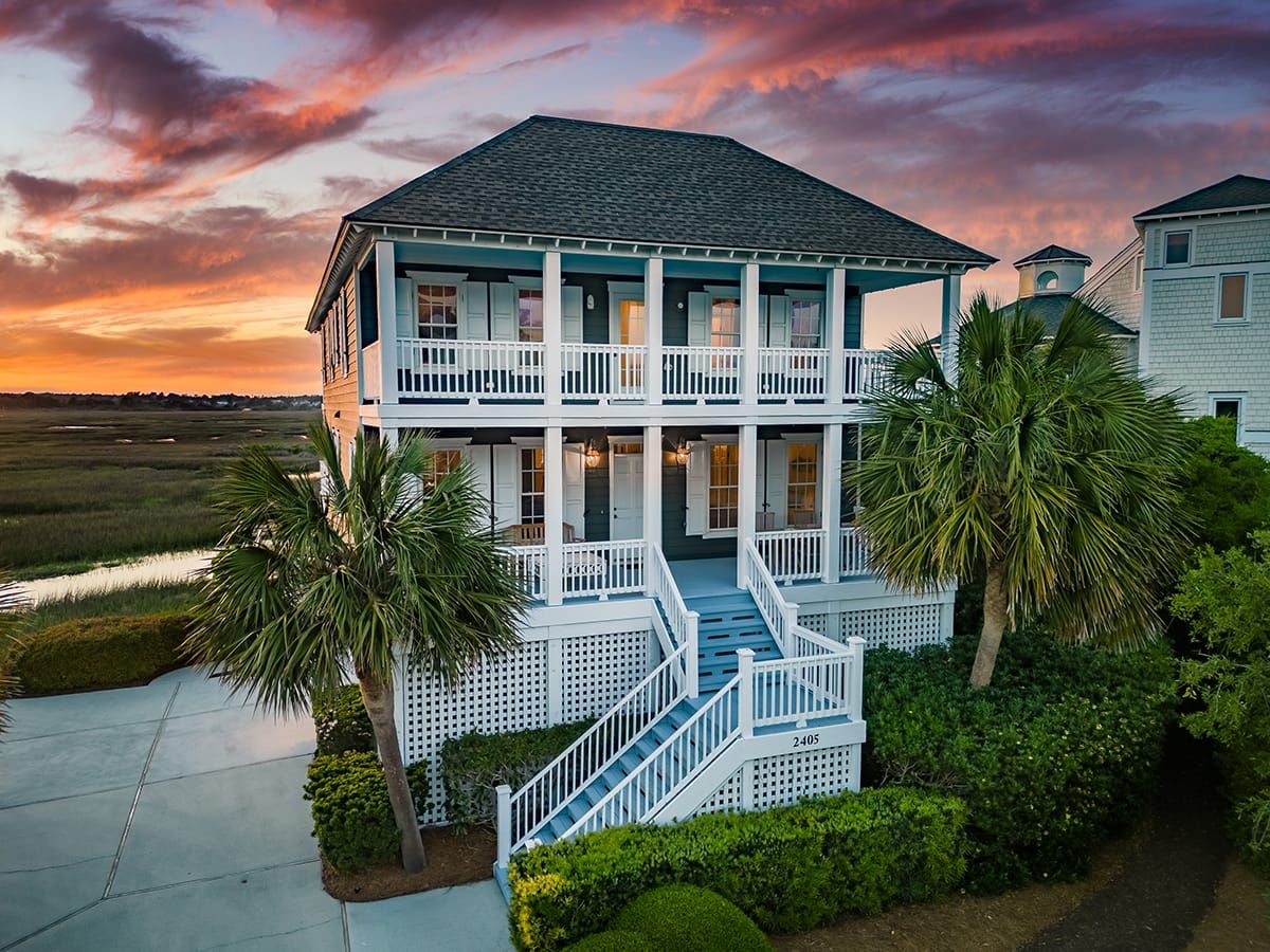 This bright Lowcountry-style home sits against a peaceful marsh with palm trees and a bold sunset sky. Perfect for big families or groups, it offers wraparound porches and plenty of space to relax after beach adventures.