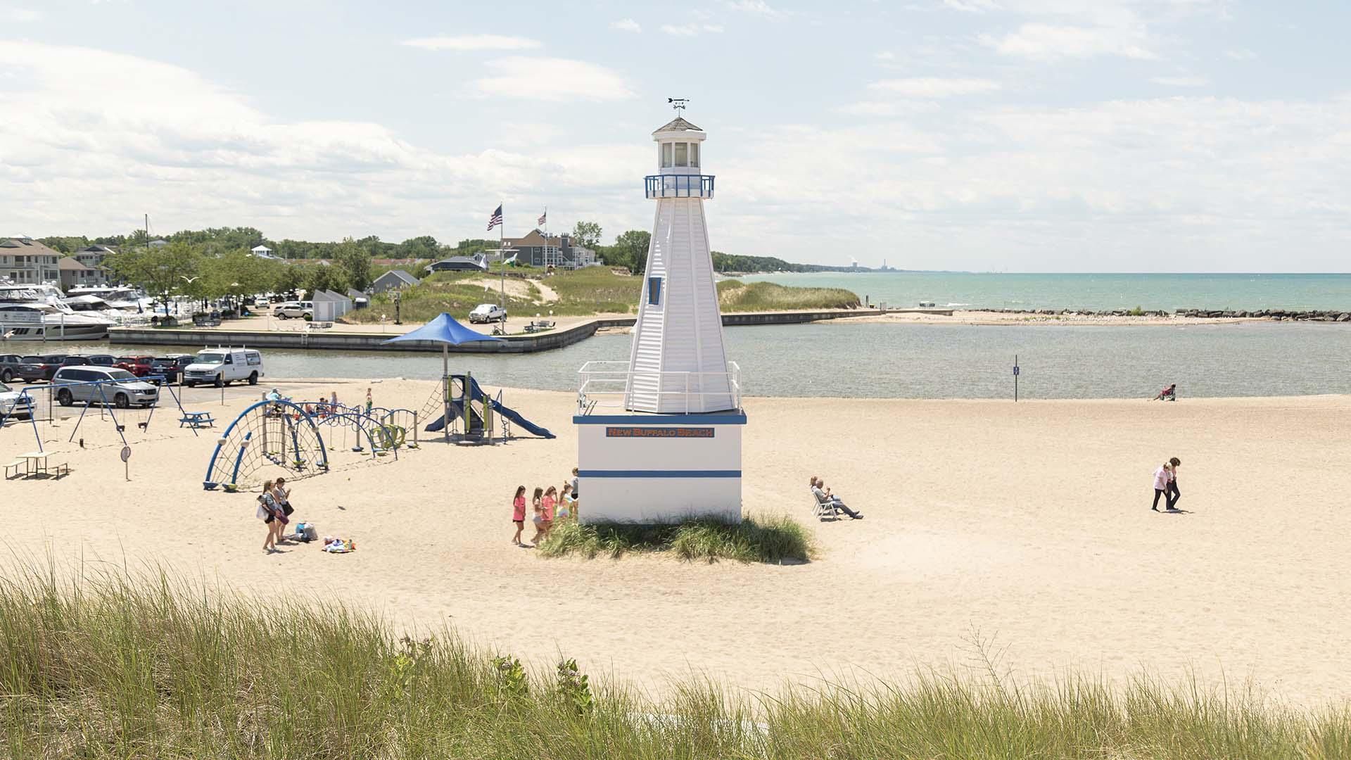 Families enjoy a sunny day at New Buffalo Beach, with children playing near the lighthouse-themed playground by the water. The soft sand, blue skies, and nearby marina create a perfect lakeside escape