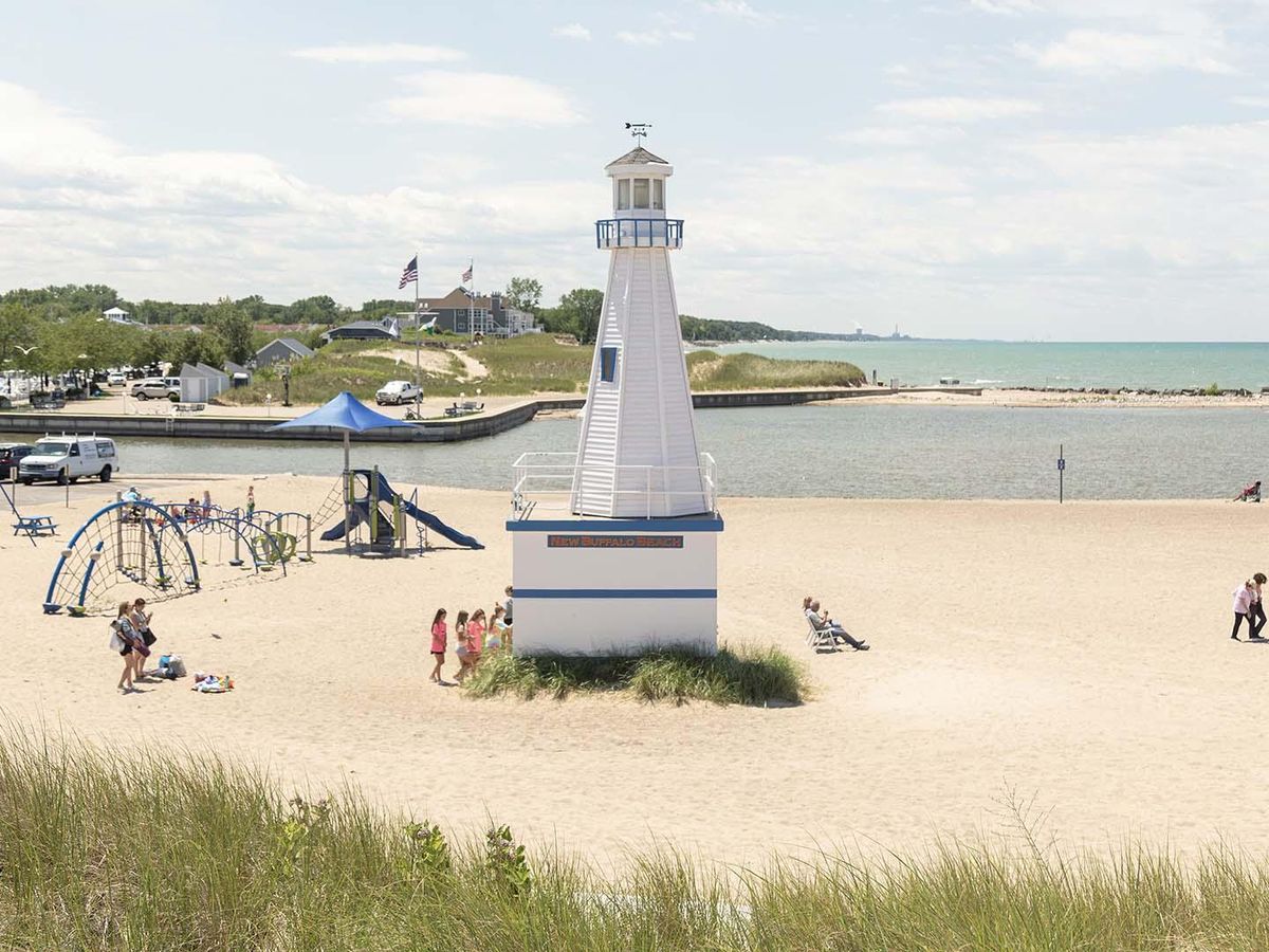 Families enjoy a sunny day at New Buffalo Beach, with children playing near the lighthouse-themed playground by the water. The soft sand, blue skies, and nearby marina create a perfect lakeside escape