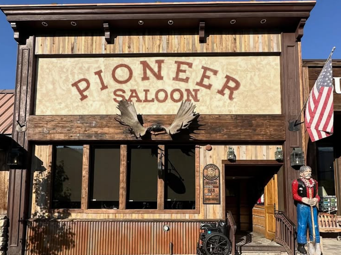 The rustic wooden facade of the Pioneer Saloon, featuring a large sign, moose antlers mounted above the entrance, an American flag, a life-size pioneer statue, and a wheelchair parked near the ramped entry.