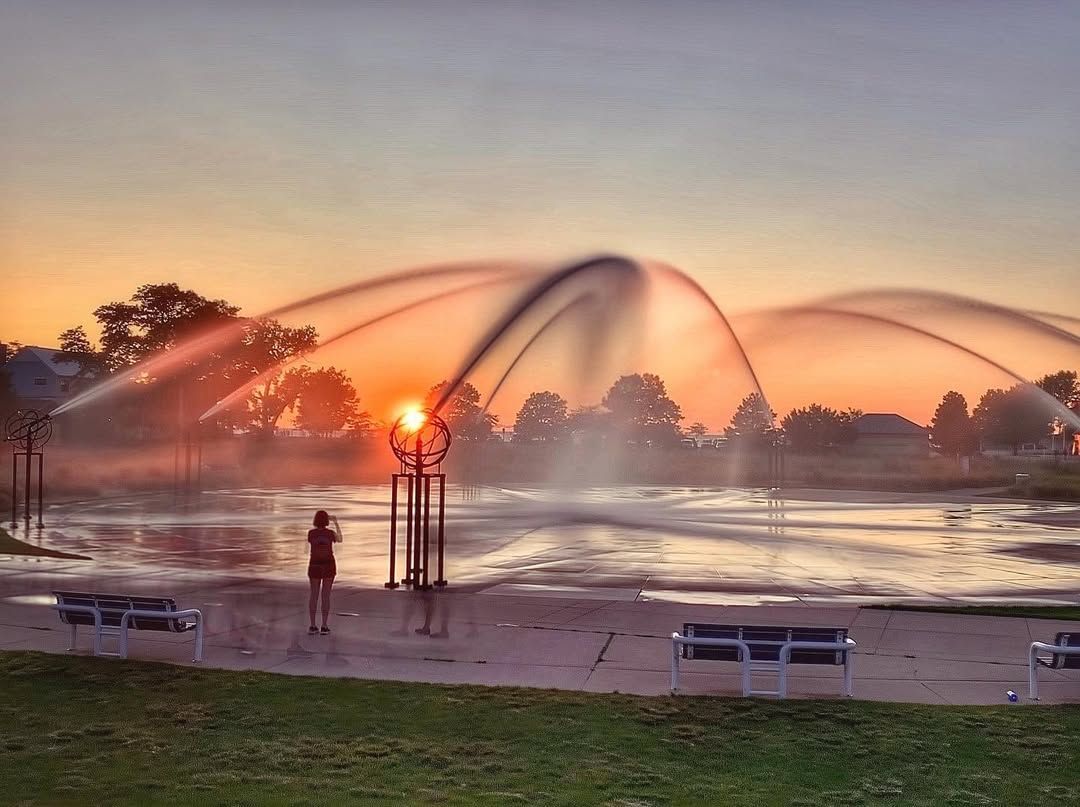 A person stands near the Whirlpool Compass Fountain at sunset as jets of water arc gracefully through the air. The warm orange glow of the setting sun creates a magical scene filled with motion and light.