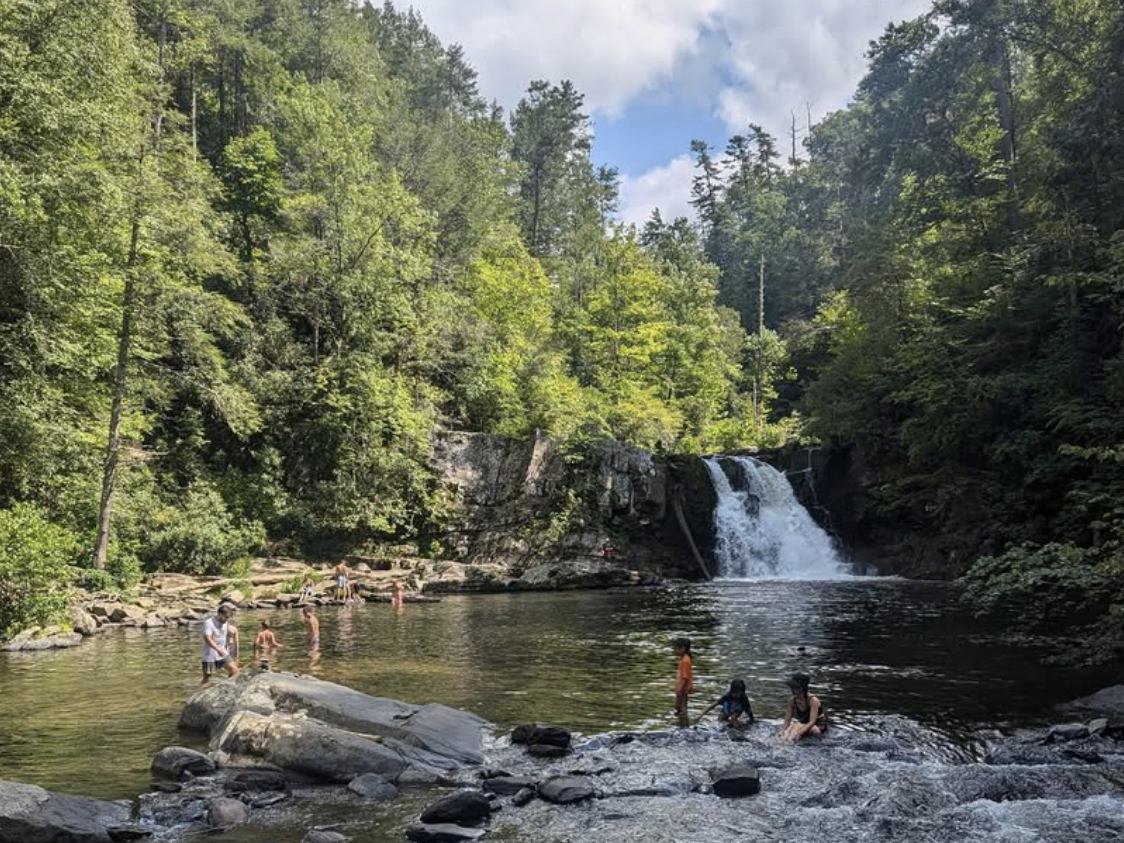 Visitors wade and relax near a waterfall surrounded by lush green forest in the Great Smoky Mountains, enjoying the natural swimming area on a sunny day.