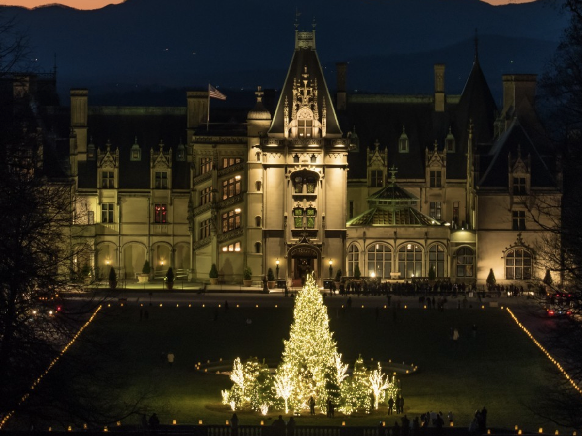 Grand historic estate illuminated at night with a large glowing Christmas tree on the front lawn, set against a backdrop of mountains at sunset