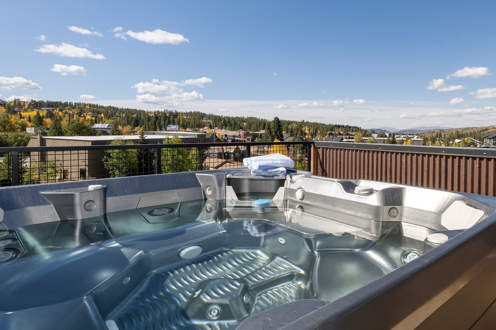 A rooftop hot tub with clear water overlooks a scenic town surrounded by trees with autumn foliage and distant mountains under a sunny blue sky.