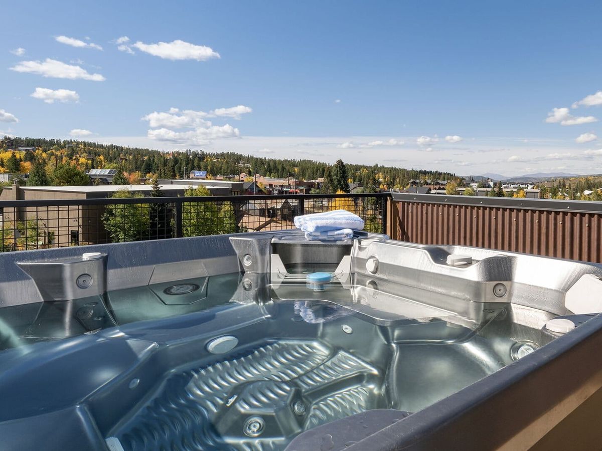 A rooftop hot tub with clear water overlooks a scenic town surrounded by trees with autumn foliage and distant mountains under a sunny blue sky.