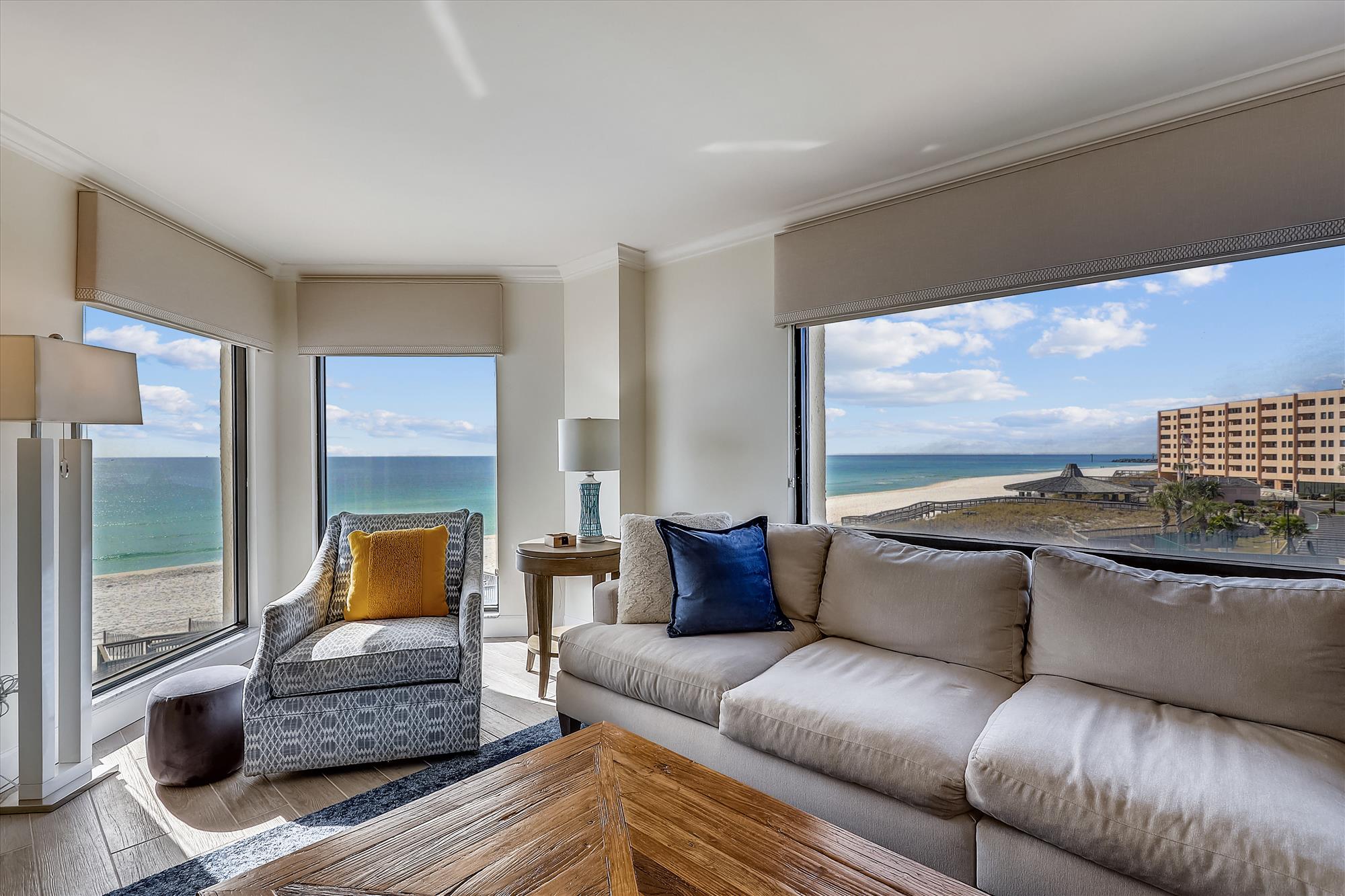 A cozy living room with panoramic beach views through large corner windows, featuring a neutral sectional sofa, patterned armchair, and a rustic wooden coffee table.