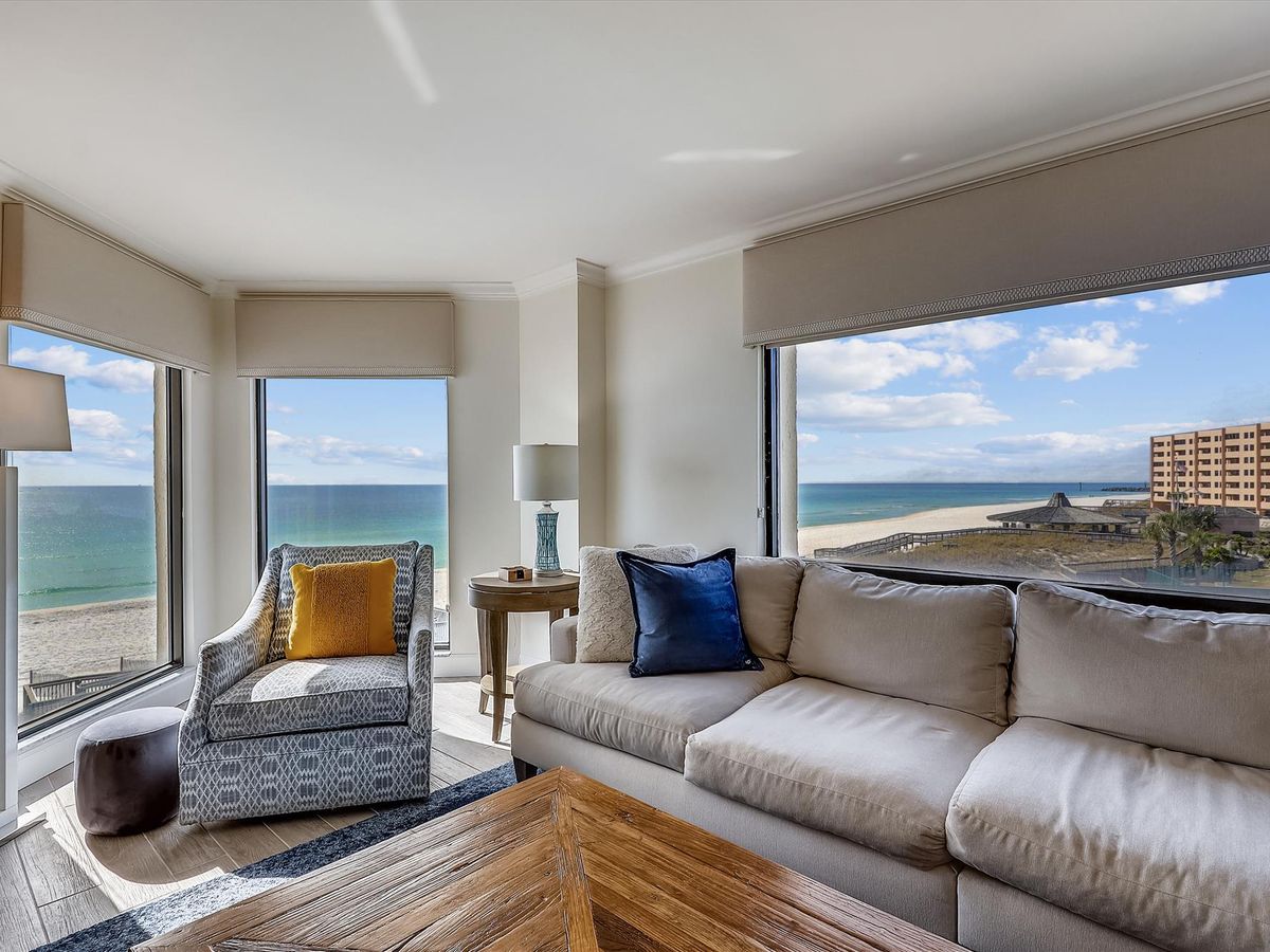 A cozy living room with panoramic beach views through large corner windows, featuring a neutral sectional sofa, patterned armchair, and a rustic wooden coffee table.