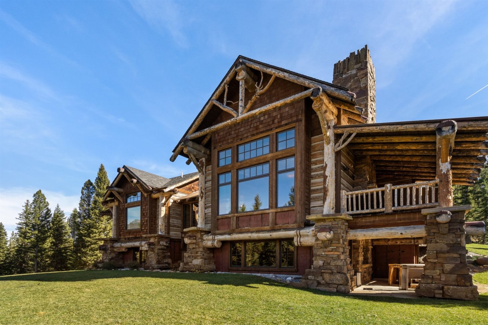 A rustic mountain lodge with large timber beams, stone accents, and expansive windows sits on a grassy lawn surrounded by pine trees under a clear blue sky.