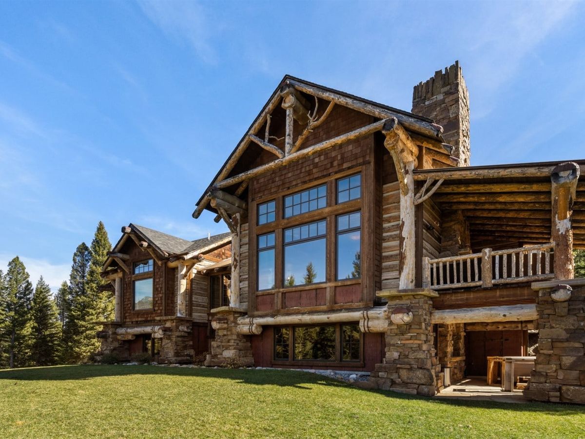 A rustic mountain lodge with large timber beams, stone accents, and expansive windows sits on a grassy lawn surrounded by pine trees under a clear blue sky.