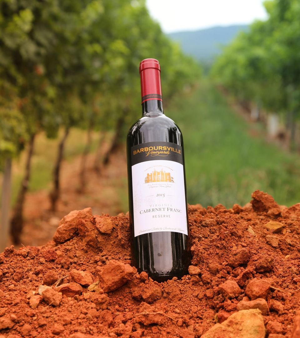 A bottle of Barboursville Vineyards Cabernet Franc rests on rich red soil between vineyard rows. The earthy tones and vineyard backdrop highlight Virginia’s fertile land and deep winemaking heritage.