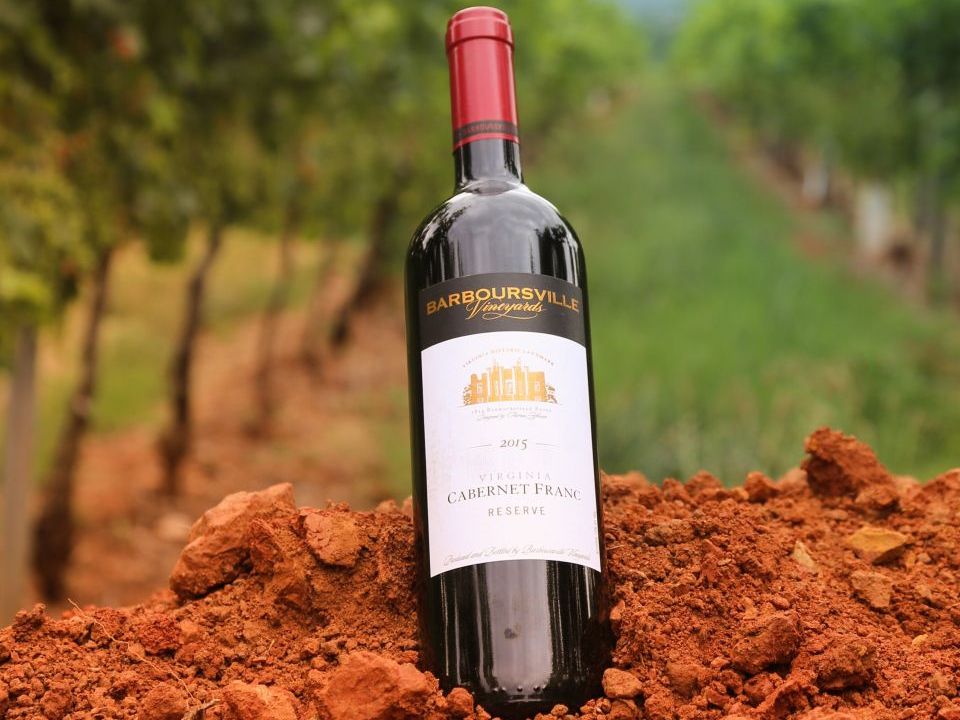 A bottle of Barboursville Vineyards Cabernet Franc rests on rich red soil between vineyard rows. The earthy tones and vineyard backdrop highlight Virginia’s fertile land and deep winemaking heritage.