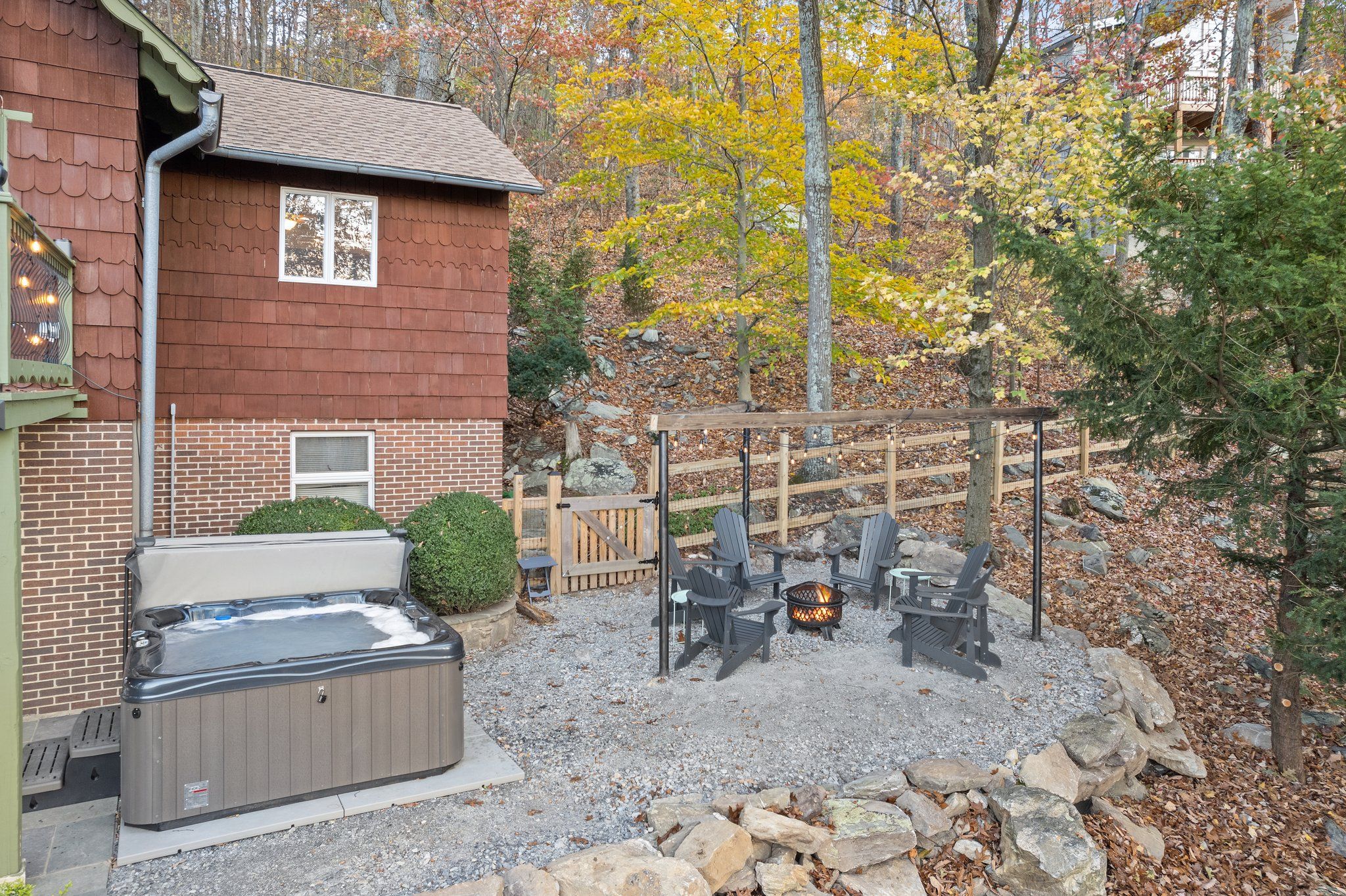 A cozy outdoor space beside a cabin featuring a covered hot tub and a gravel fire pit area with Adirondack chairs, surrounded by autumn trees and a rustic wooden fence.