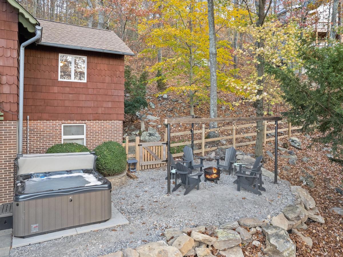 A cozy outdoor space beside a cabin featuring a covered hot tub and a gravel fire pit area with Adirondack chairs, surrounded by autumn trees and a rustic wooden fence.