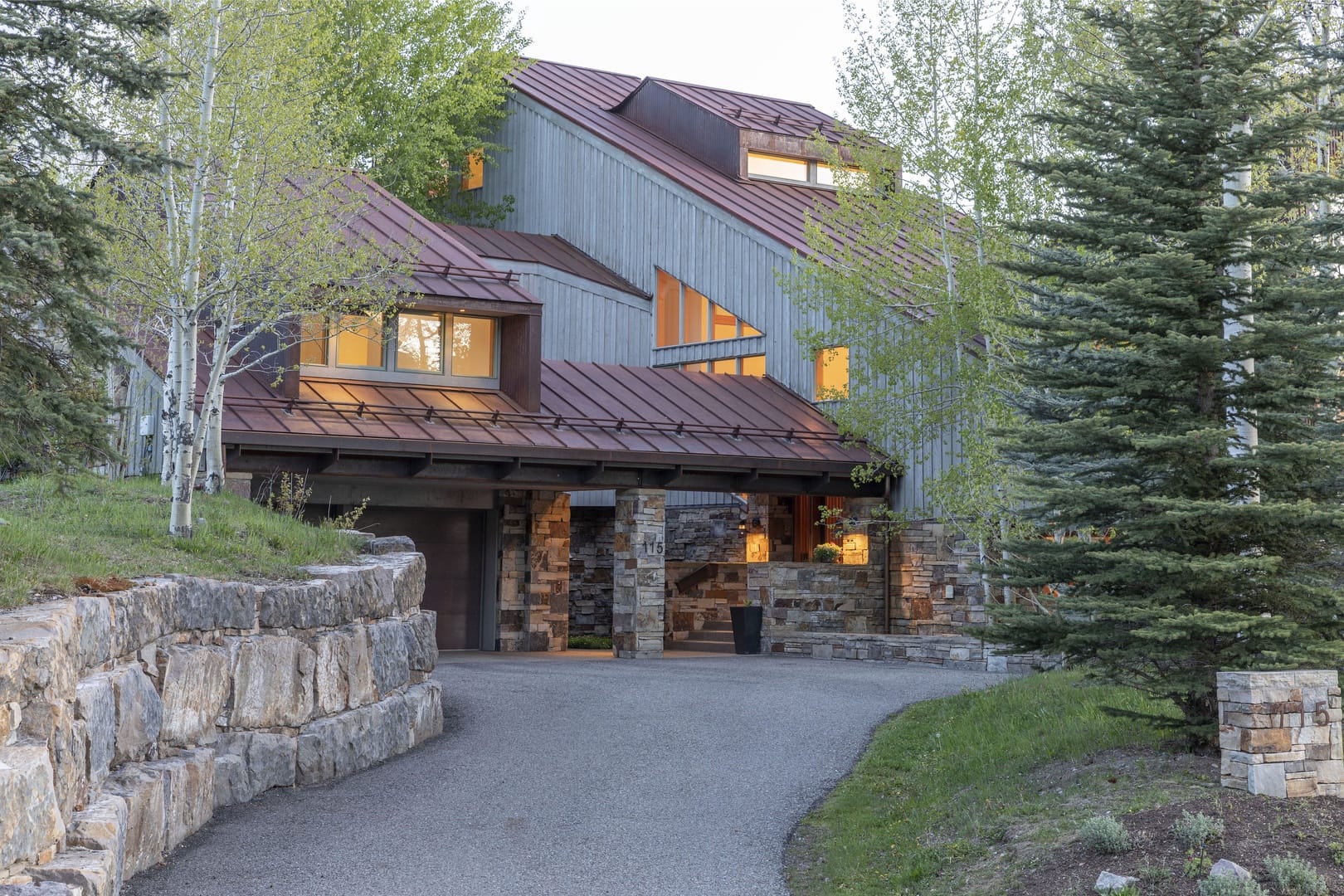 A contemporary mountain home with metal roofing, vertical wood siding, and stone accents, nestled among aspens and evergreens with warm interior lights glowing at dusk.