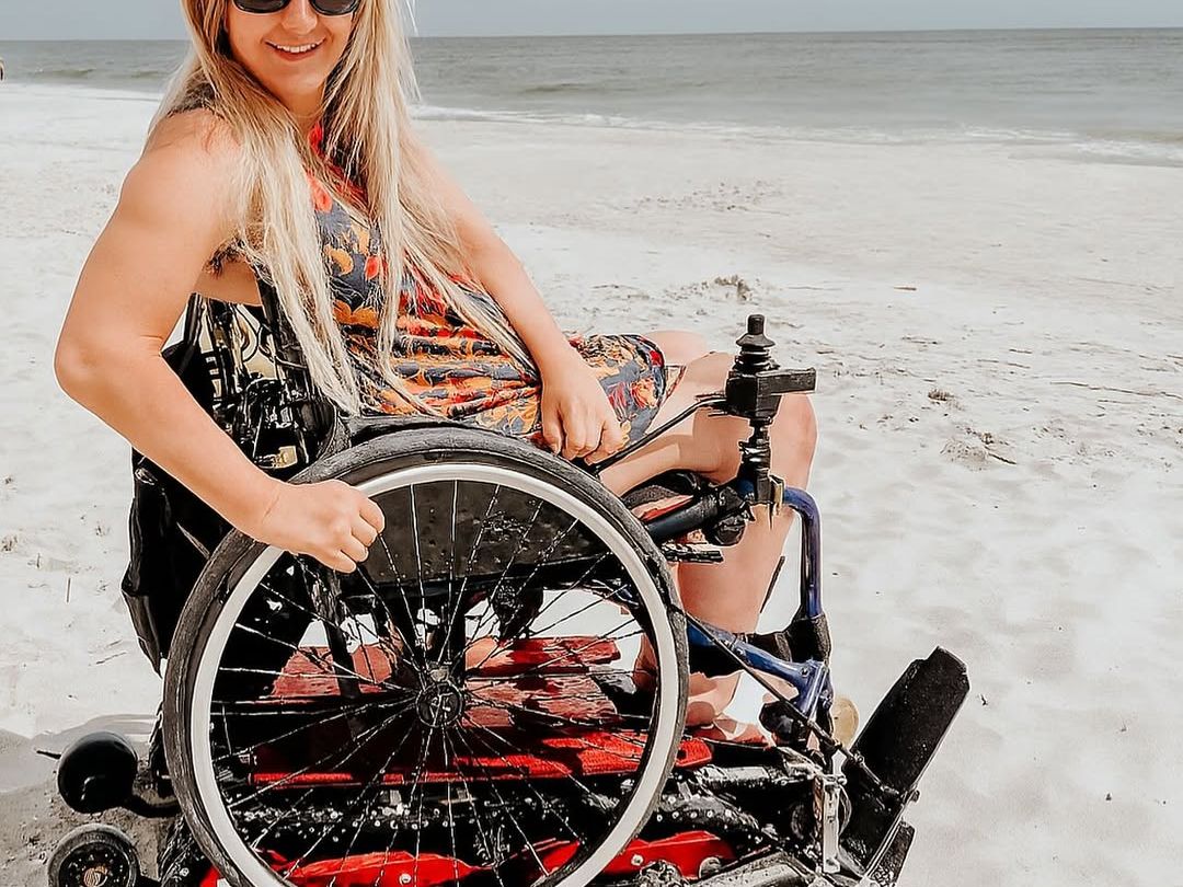 A woman enjoys a sunny day at the beach in a fat tire wheelchair, designed for smooth movement across the sand. Her smile reflects the freedom and accessibility this adaptive rental provides.