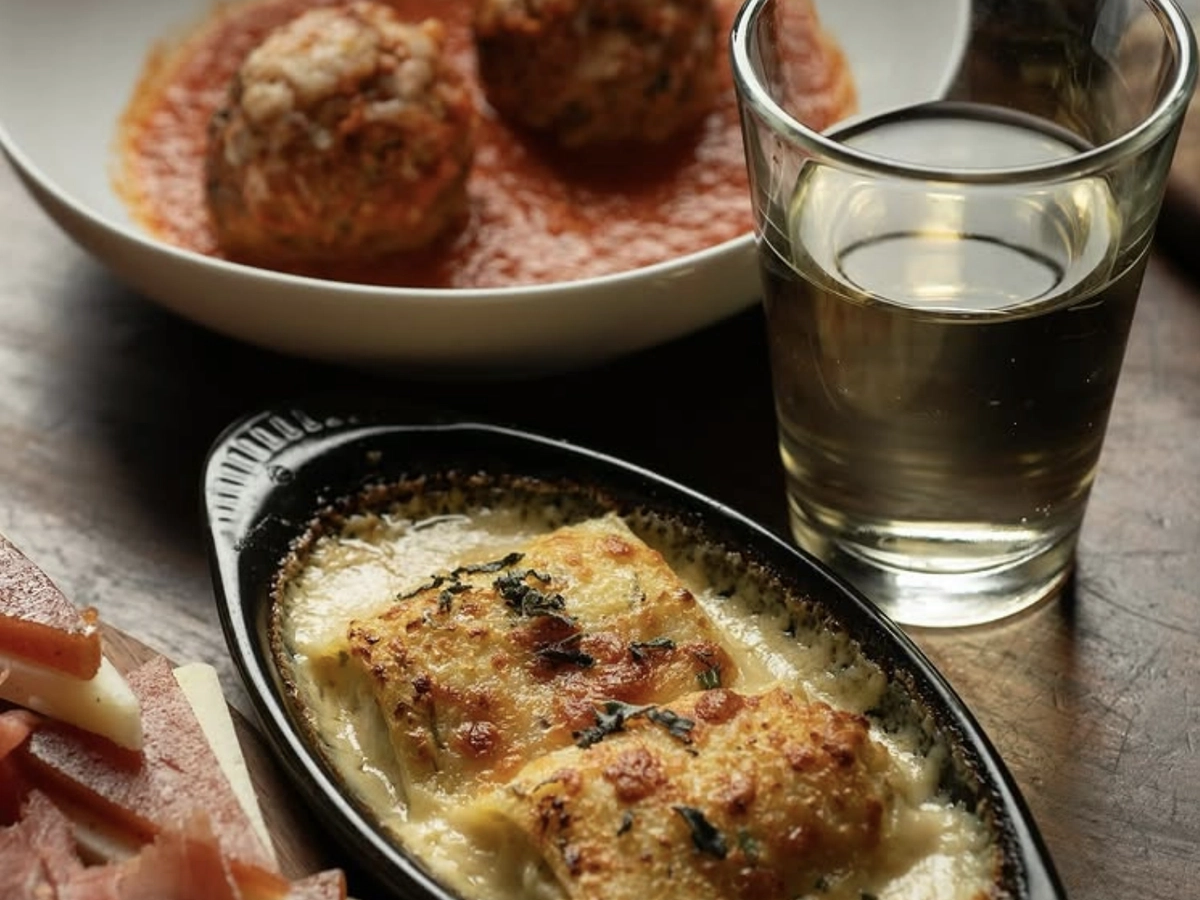 Close-up of an Italian meal featuring baked pasta in a creamy sauce, meatballs in tomato sauce, sliced cheese with cured meat, and a glass of white wine.