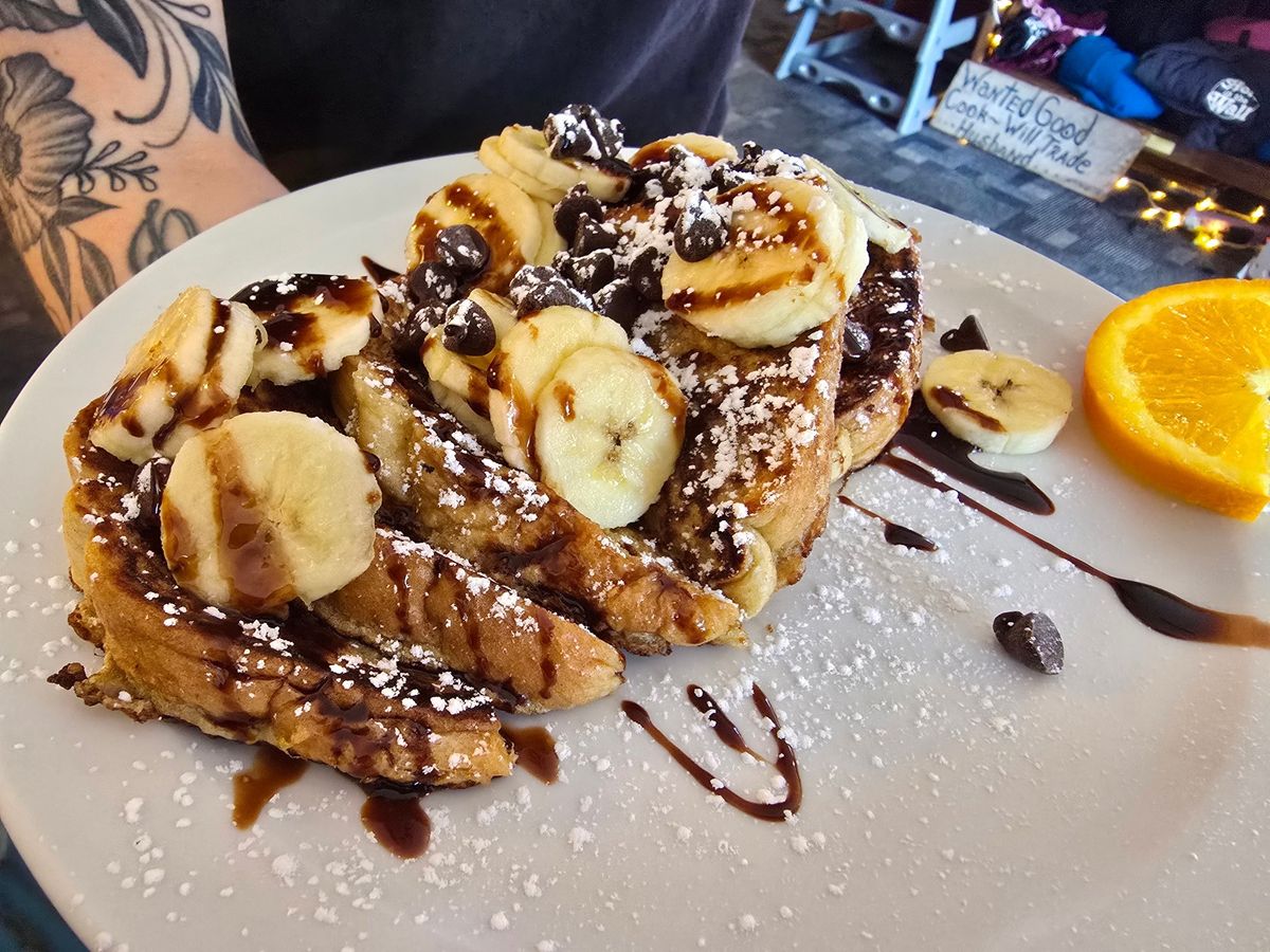 Thick slices of French toast are piled high and topped with fresh banana slices, chocolate chips, a dusting of powdered sugar, and a drizzle of chocolate sauce, served alongside an orange wedge. A tattooed arm is visible in the background beside a handwritten chalkboard sign, adding to the casual, colorful atmosphere.