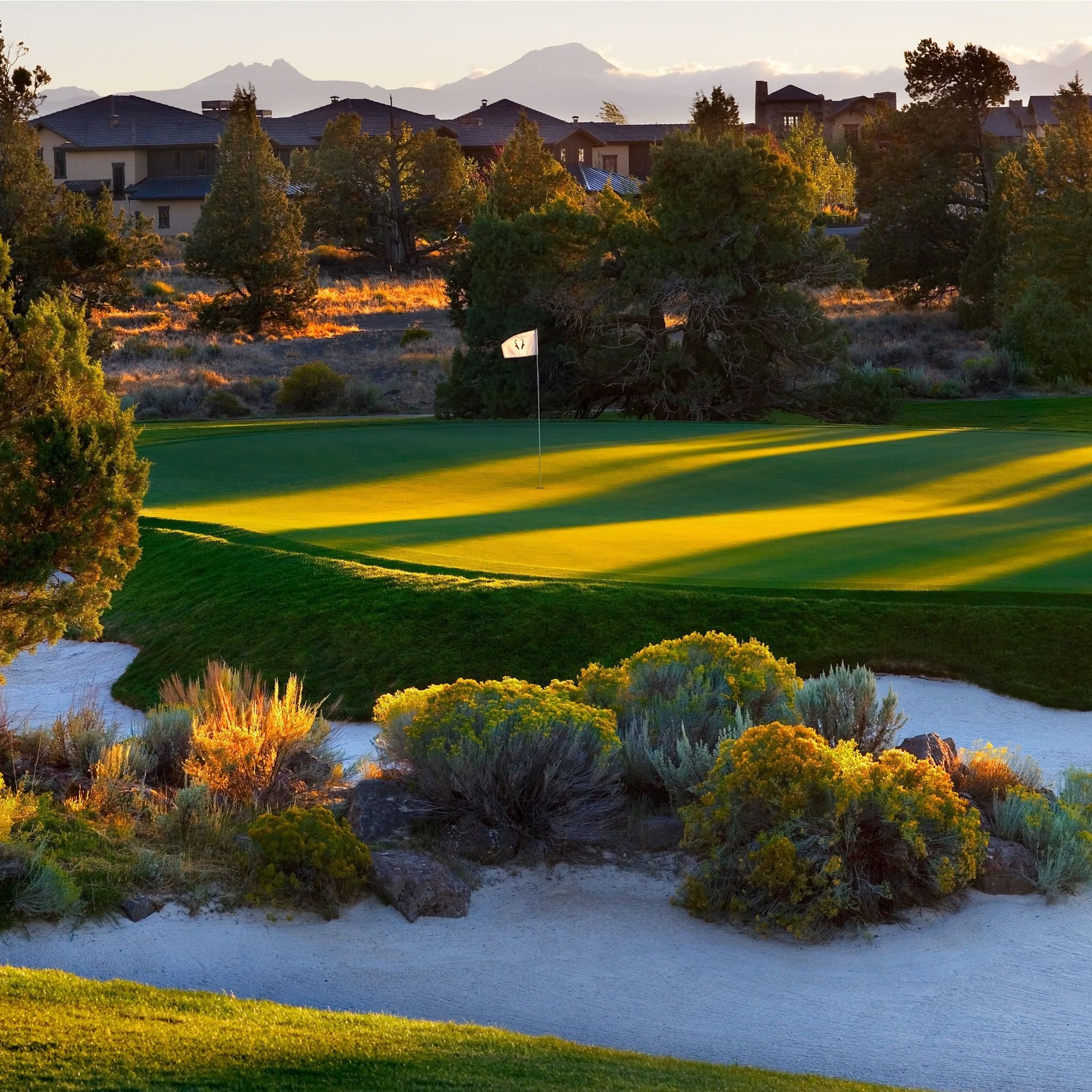 Late afternoon golden light casts long shadows across a well-bunkered green at Pronghorn's Nicklaus Course in Bend, Oregon, surrounded by sagebrush, juniper trees, and resort residences against a mountain backdrop.