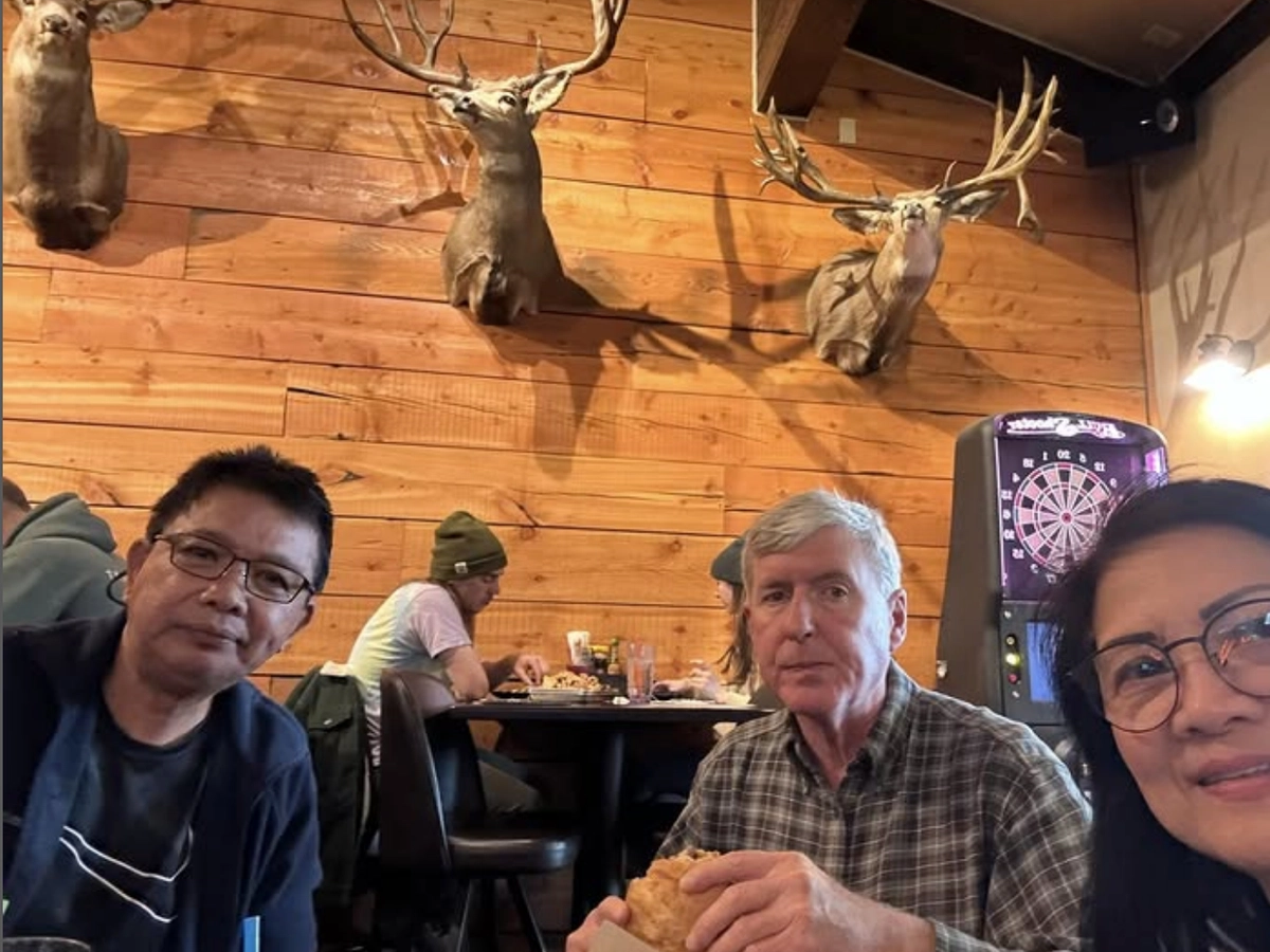 Three people enjoying a meal inside a rustic restaurant with wood-paneled walls, mounted deer heads, and a dartboard in the background.