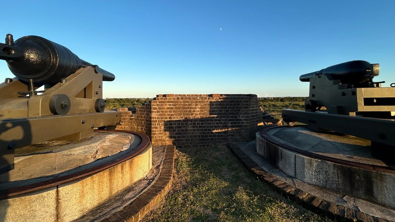 Two historic cannons sit atop the brick walls of Fort Pulaski, facing out toward the horizon. The golden sunlight highlights the fort’s preserved details and its sweeping views of the surrounding marshlands.