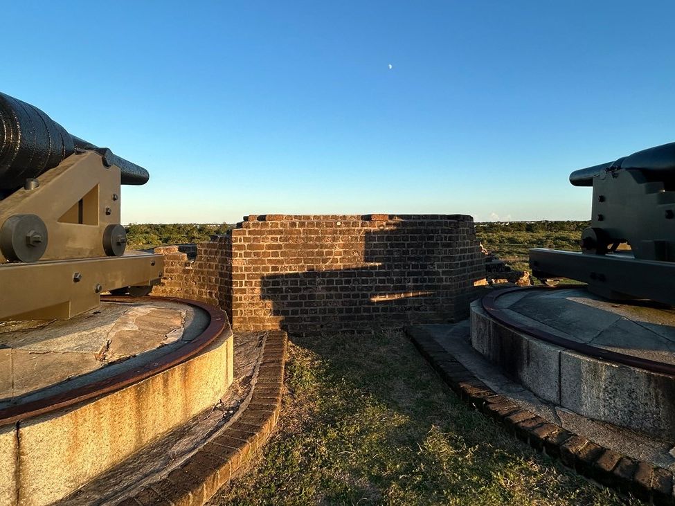 Two historic cannons sit atop the brick walls of Fort Pulaski, facing out toward the horizon. The golden sunlight highlights the fort’s preserved details and its sweeping views of the surrounding marshlands.