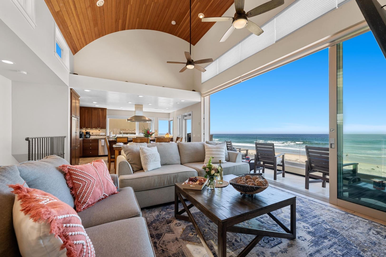 Sunlit beachfront living room with vaulted wood ceilings, neutral sofas, and large sliding glass doors opening to an oceanfront patio with uninterrupted sea views.