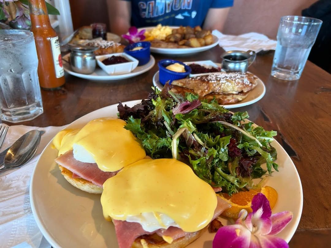 Classic eggs benedict with ham and hollandaise sauce, paired with a fresh green salad, pancakes, and hearty breakfast sides in a cozy cafe setting.