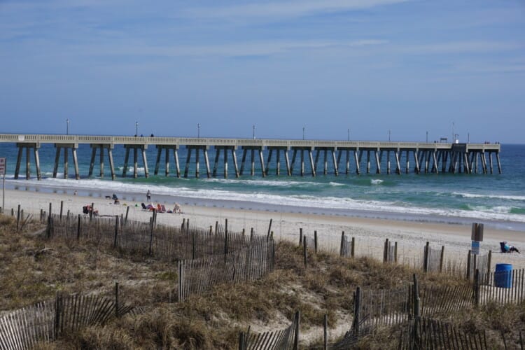 A long fishing pier reaches into the ocean at Wrightsville Beach, North Carolina, with dunes and sea oats in the foreground. Small groups of people sit along the sandy shore near the water. Wrightsville Beach offers scenic coastal views and classic Carolina beach charm.