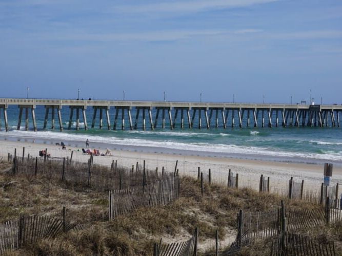 A long fishing pier reaches into the ocean at Wrightsville Beach, North Carolina, with dunes and sea oats in the foreground. Small groups of people sit along the sandy shore near the water. Wrightsville Beach offers scenic coastal views and classic Carolina beach charm.