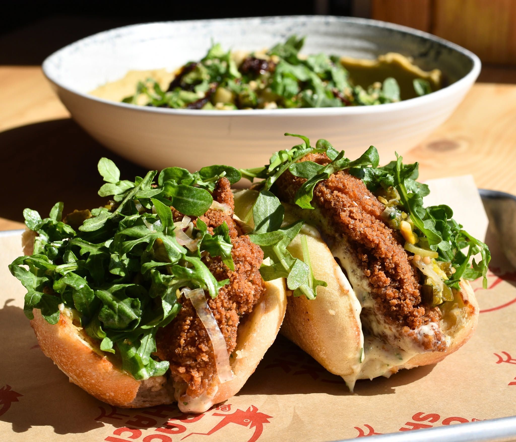 Two crispy fried chicken sandwiches piled high with fresh arugula and tangy slaw sit on a paper-lined tray, their golden cutlets peeking from soft toasted buns. A large salad bowl rests in the sunlit background at the casual eatery.