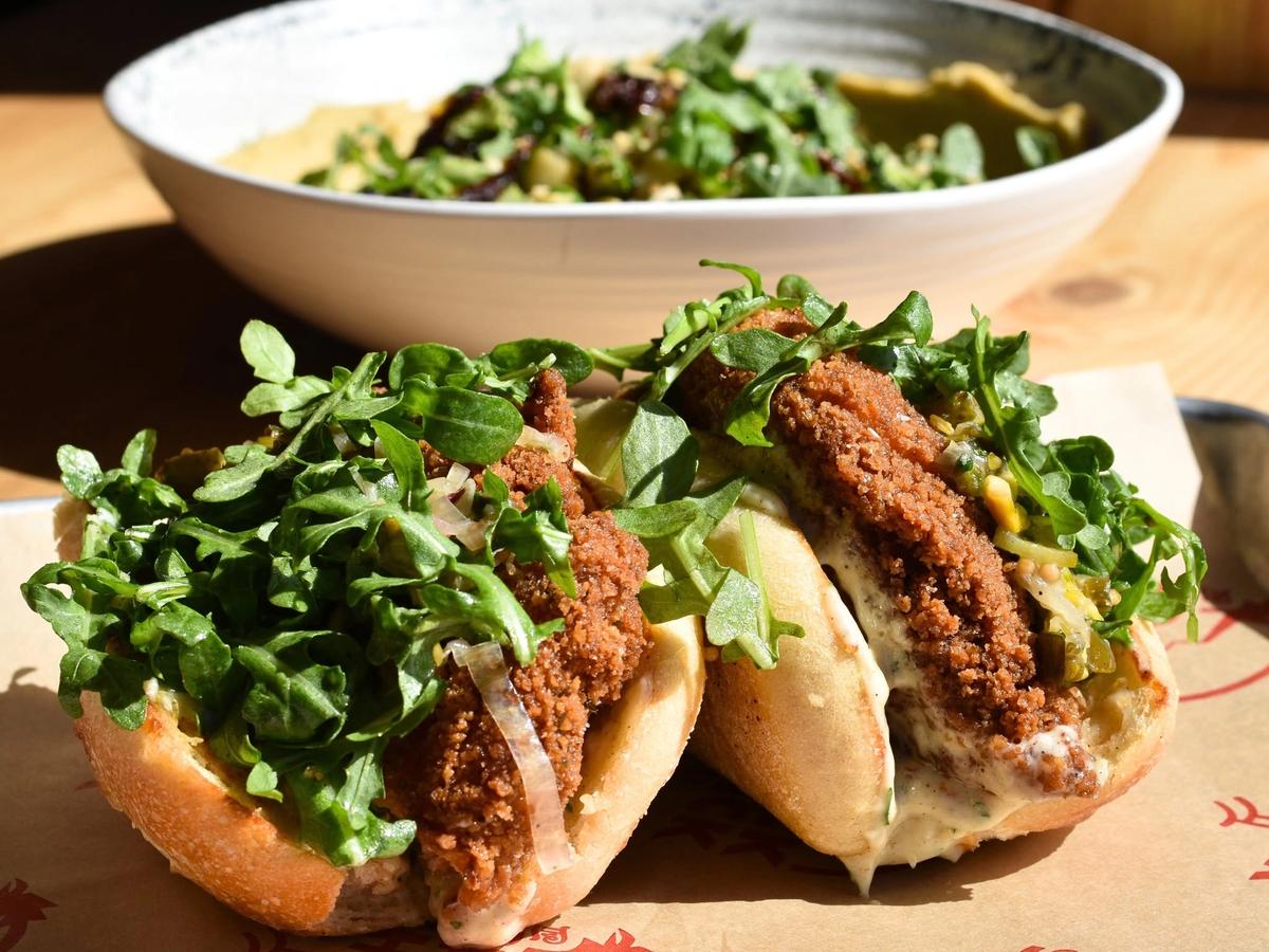 Two crispy fried chicken sandwiches piled high with fresh arugula and tangy slaw sit on a paper-lined tray, their golden cutlets peeking from soft toasted buns. A large salad bowl rests in the sunlit background at the casual eatery.