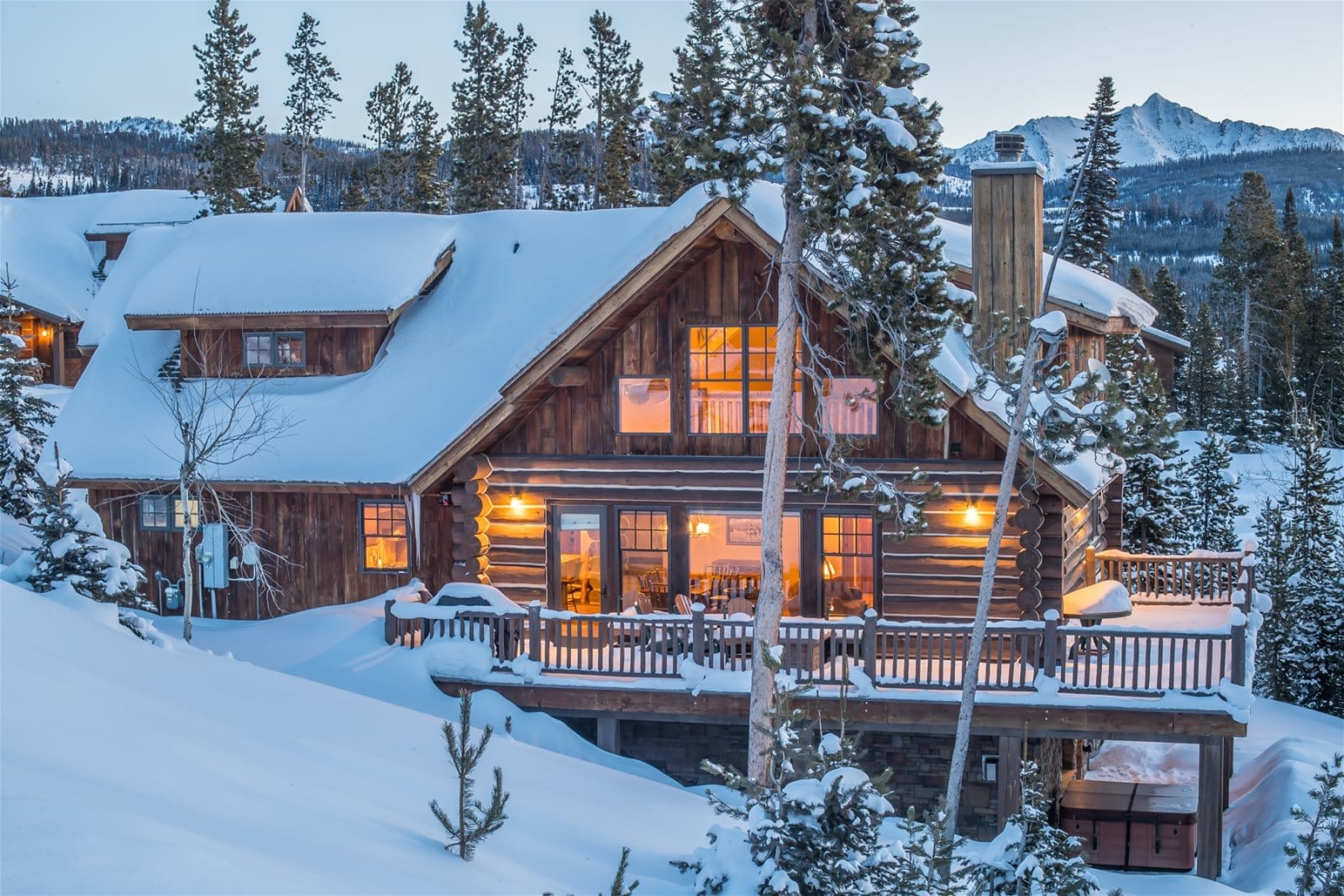 A rustic log cabin glows warmly from within as it sits nestled in deep snow, surrounded by pine trees and mountain peaks at dusk.