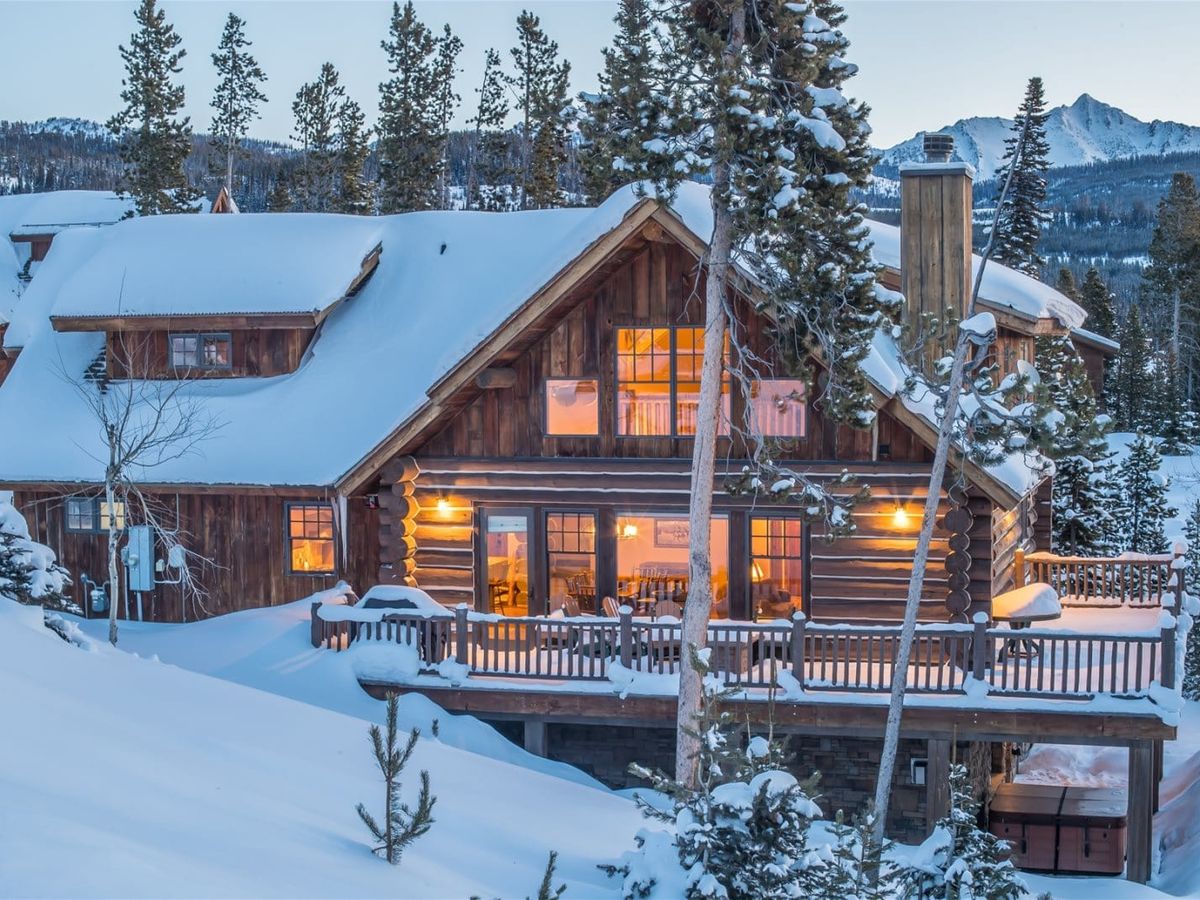 A rustic log cabin glows warmly from within as it sits nestled in deep snow, surrounded by pine trees and mountain peaks at dusk.