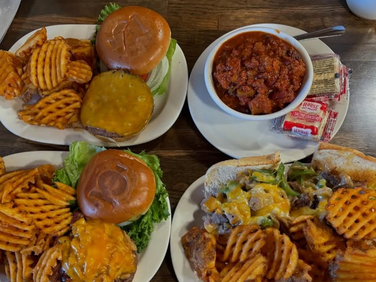 Three plates of burgers and waffle fries, a cheesy Philly cheesesteak sandwich, and a bowl of hearty chili with saltine crackers on a wooden table.