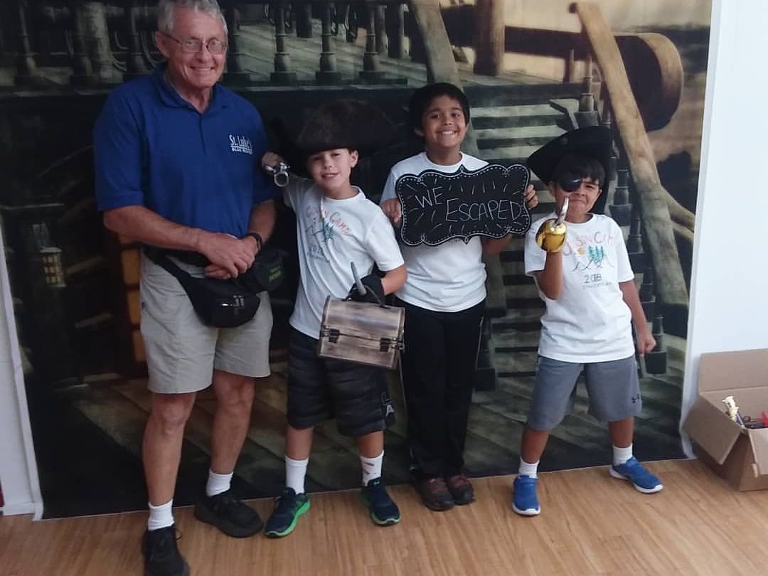 A family group poses after completing a pirate-themed escape room, smiling with props like hats, swords, and treasure chests. The sign “We Escaped” shows off their success. The playful setting creates a fun memory for both kids and adults.