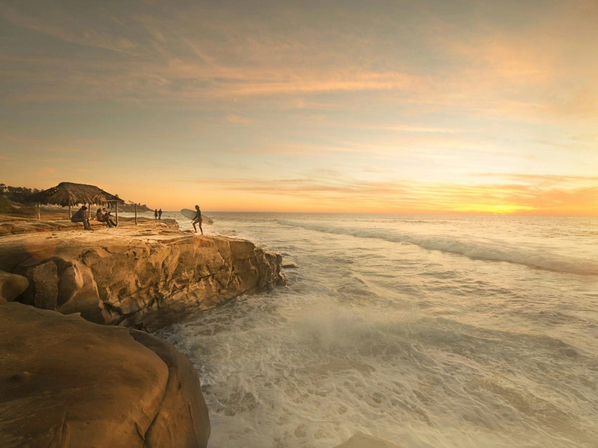 A surfer holding a board stands on a rocky shoreline at sunset as waves crash below and people relax under a thatched hut nearby.