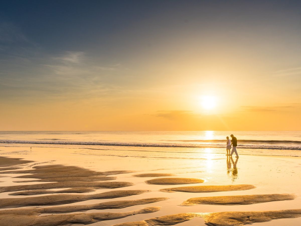 A serene beach scene at sunrise, with a couple walking along the shoreline. The golden light reflects off the wet sand and tide pools, creating a peaceful and romantic atmosphere