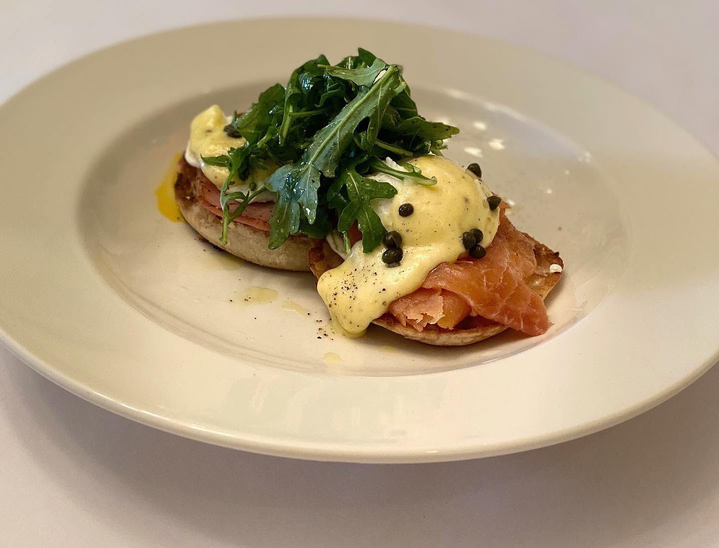 An elegant eggs Benedict rests on a white plate, layering a toasted English muffin with smoked salmon, poached eggs, and rich hollandaise dotted with capers. A bright tangle of dressed arugula tops the dish, with golden yolk pooling on the plate.